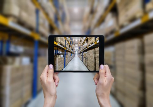 Horizontal color image of female hands holding a digital tablet in a corridor of futuristic distribution warehouse. Ordering online from a modern warehouse on a touchscreen tablet computer. Large distribution storage in background with racks full of packages, boxes, pallets, crates ready to be delivered. Logistics, freight, shipping, receiving.